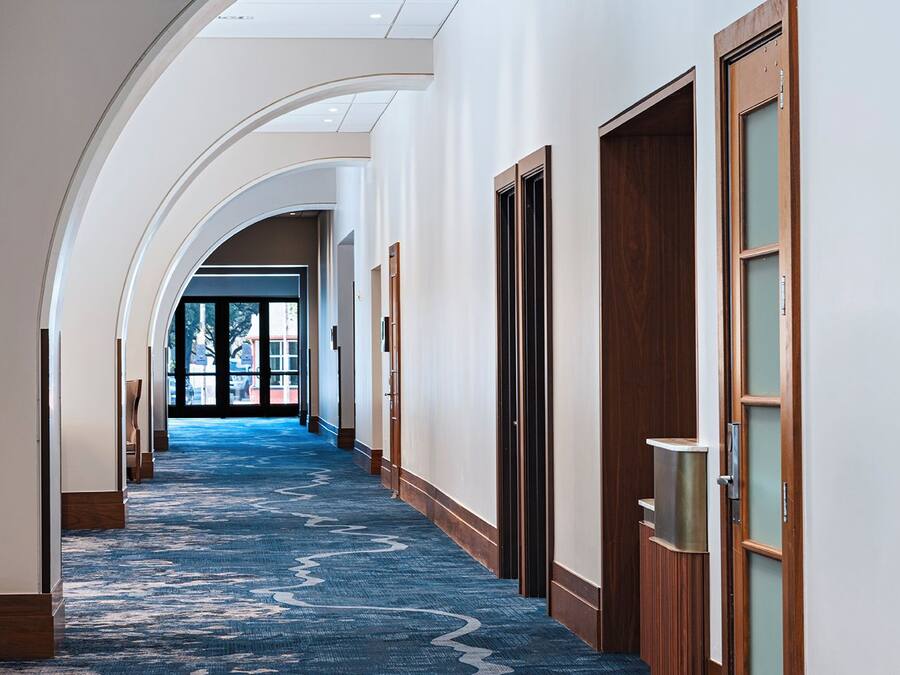 A hotel corridor with a blue carpet, wooden doors, and a skylight.