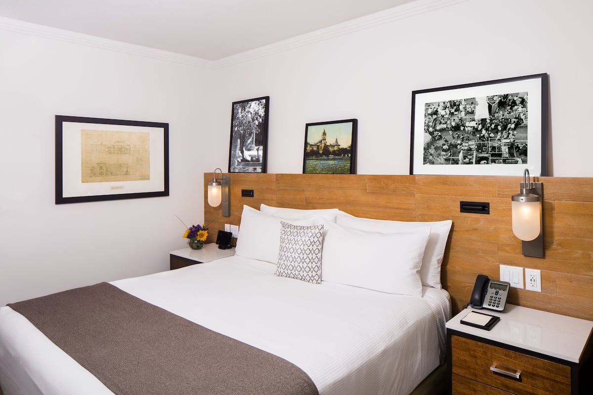 Habitación de hotel con una cama grande, cabecera de madera, mesita de noche con lámpara y cuadros enmarcados en la pared.