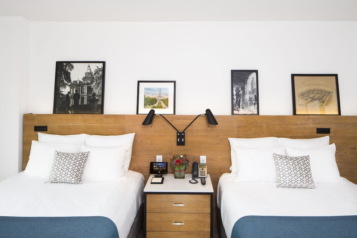 Habitación de hotel con dos camas, cabecera de madera, mesita de noche con un teléfono, una pequeña mesa con un florero y cuadros en la pared.