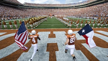 A football stadium with a marching band and players running onto the field.