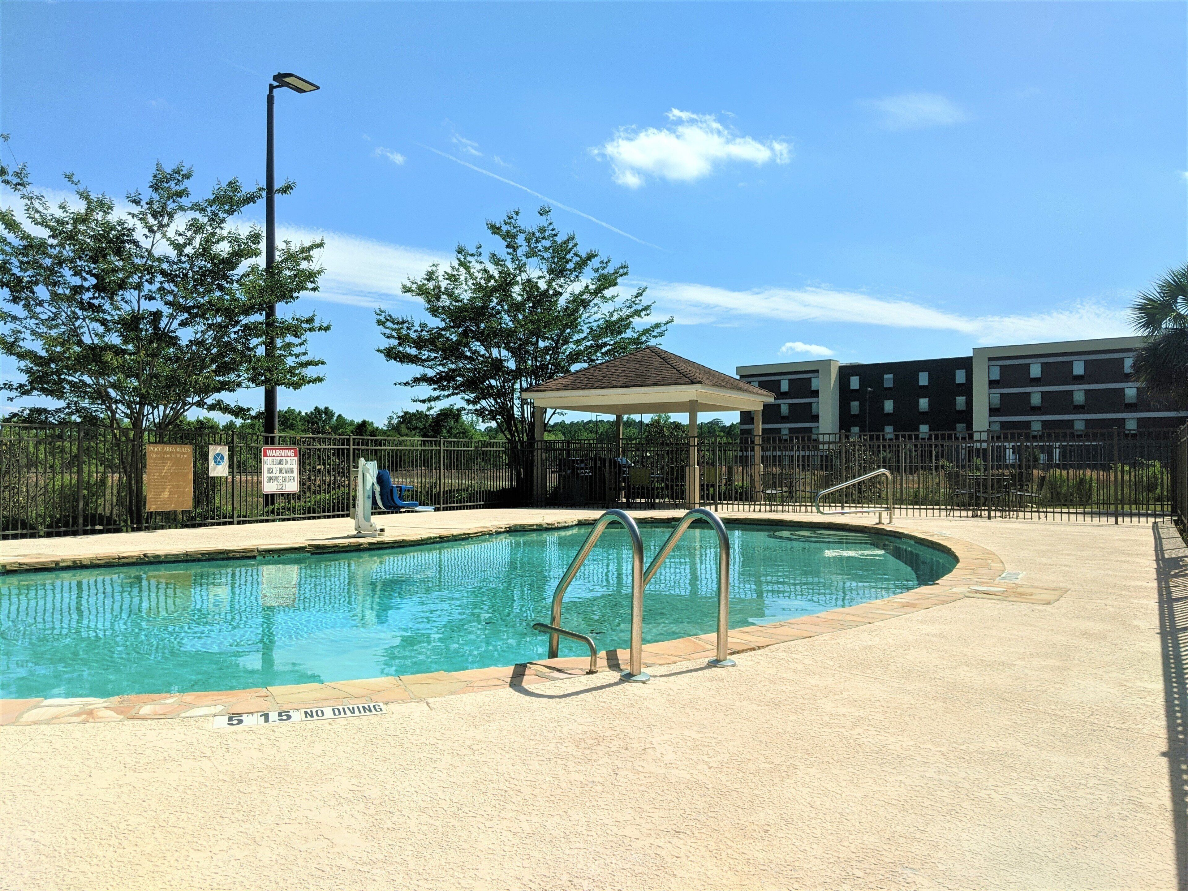 A swimming pool with a no diving sign, a covered seating area, and a fenced enclosure with trees and a building in the background.