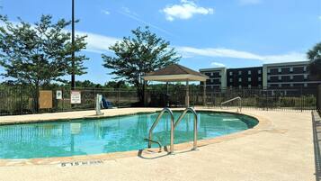 A swimming pool with a no diving sign, a covered seating area, and a fenced enclosure with trees and a building in the background.