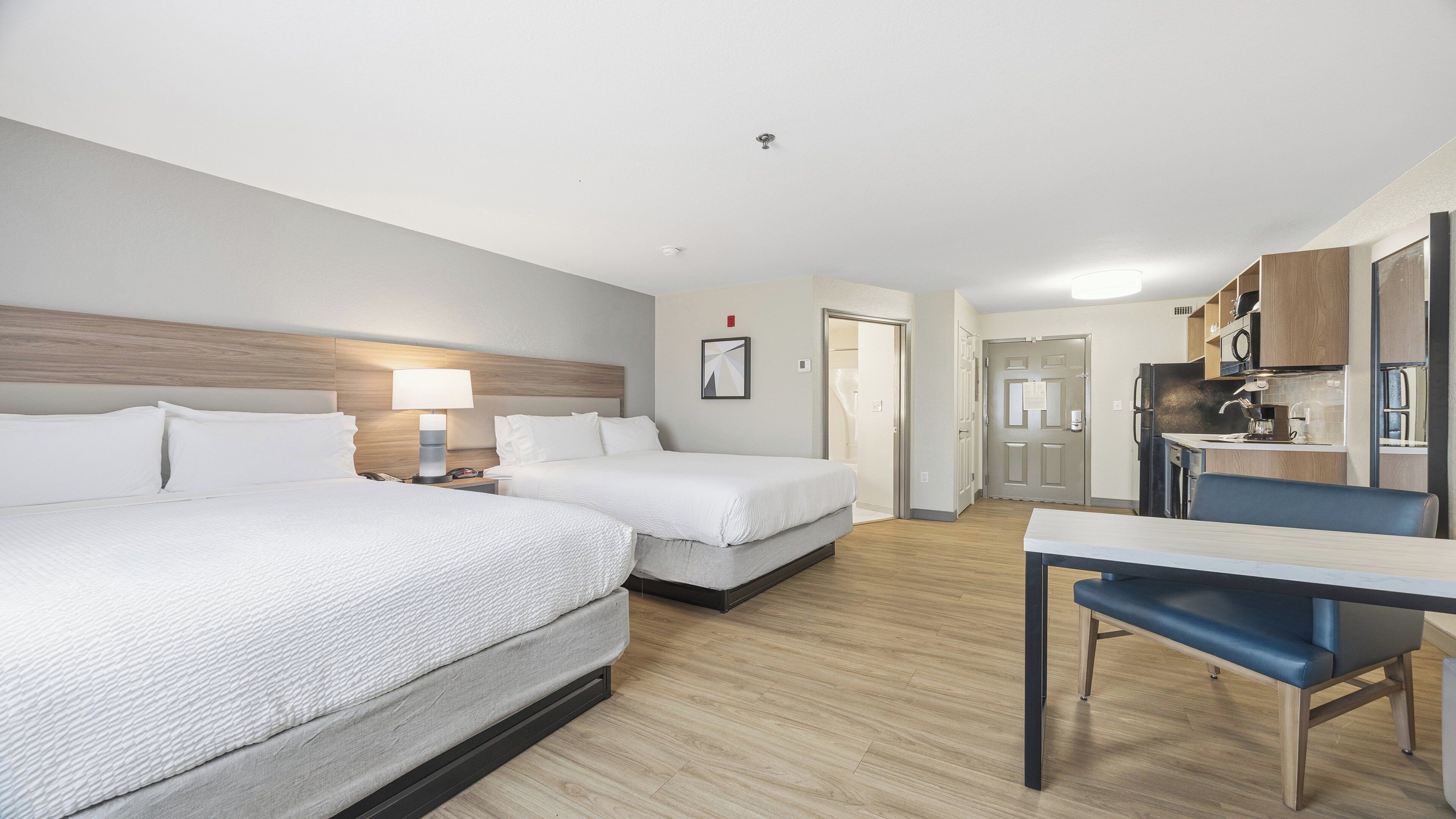 A hotel room with two beds, a desk, a chair, a kitchenette, and a bathroom.