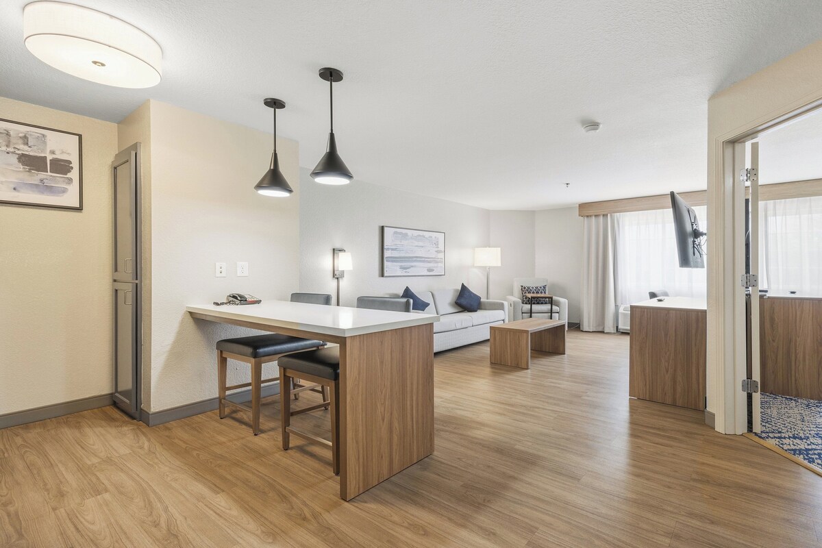 Una habitación de hotel moderna con kitchenette, zona de comedor, sof á, televisor y un ventanal grande.