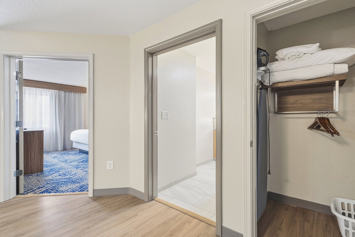 Habitación de hotel con cama, baño con ducha, suelo de madera y un cesto de ropa.