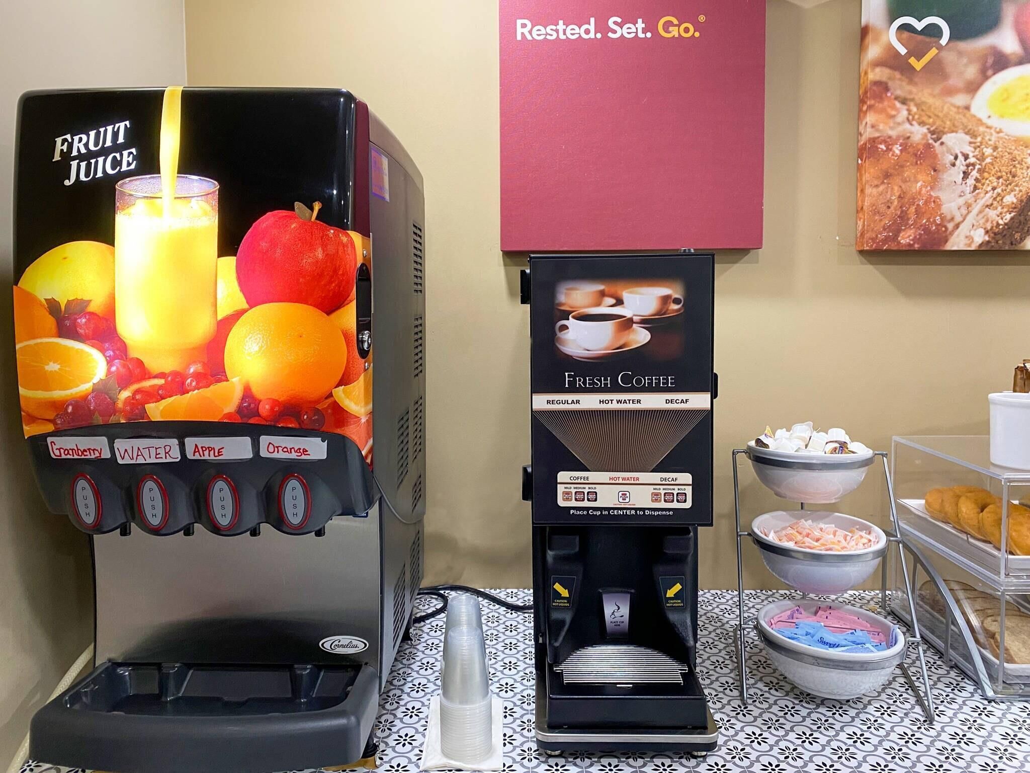 Un dispensador de jugo con opciones de fruta y una máquina de café con variedad de bebidas.