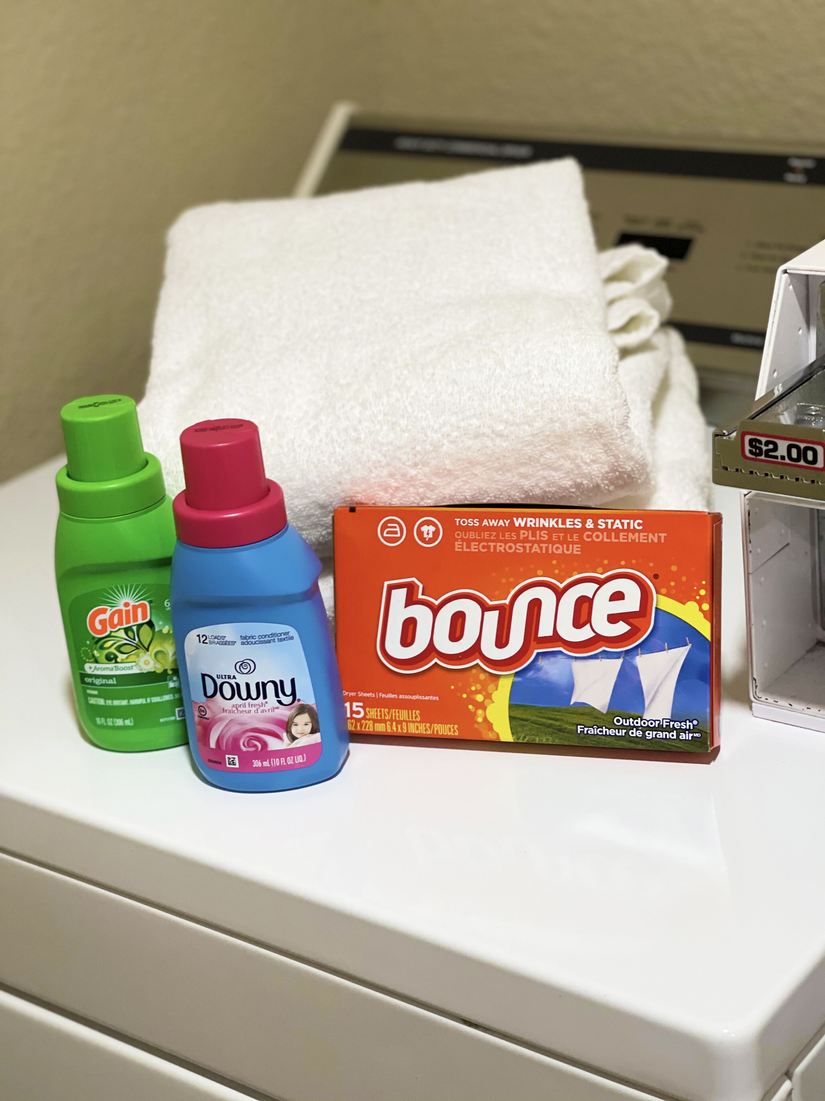 Una cesta de ropa con una toalla blanca, detergente Gain, suavizante Downy y hojas de secadora Bounce.