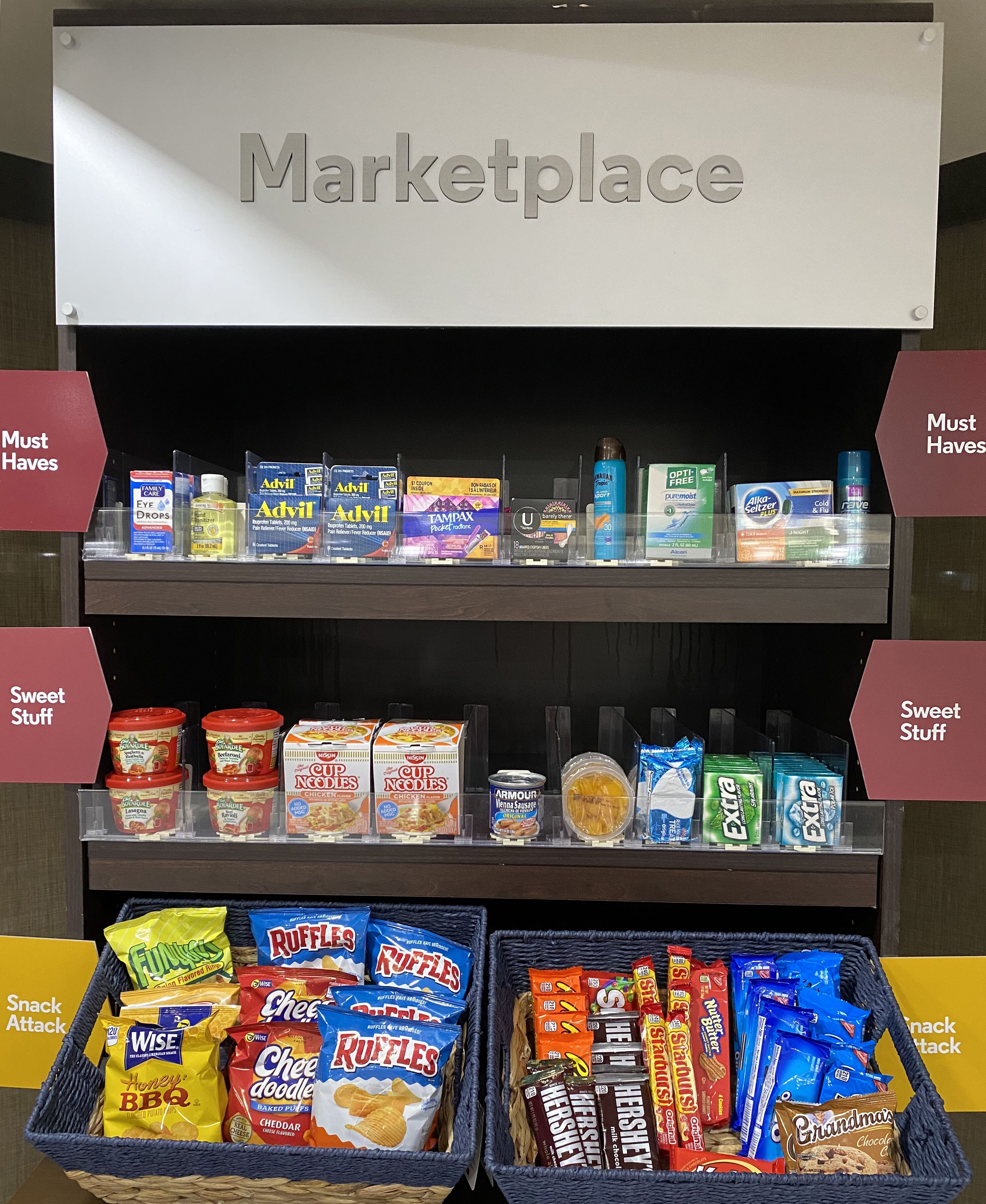 Una presentación en tienda con estantes etiquetados 'Imprescindibles' y 'Delicias', mostrando productos como Advil, Tampax y diversos snacks.