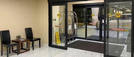 Entrada de hotel con puerta de cristal, señal de precaución, extintor y rampa accesible para sillas de ruedas.