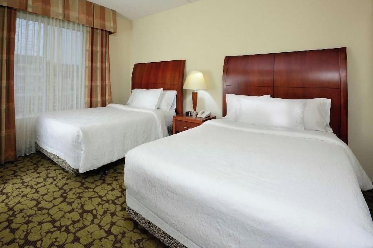 A hotel room with two beds, a nightstand with a lamp, and a window with curtains.