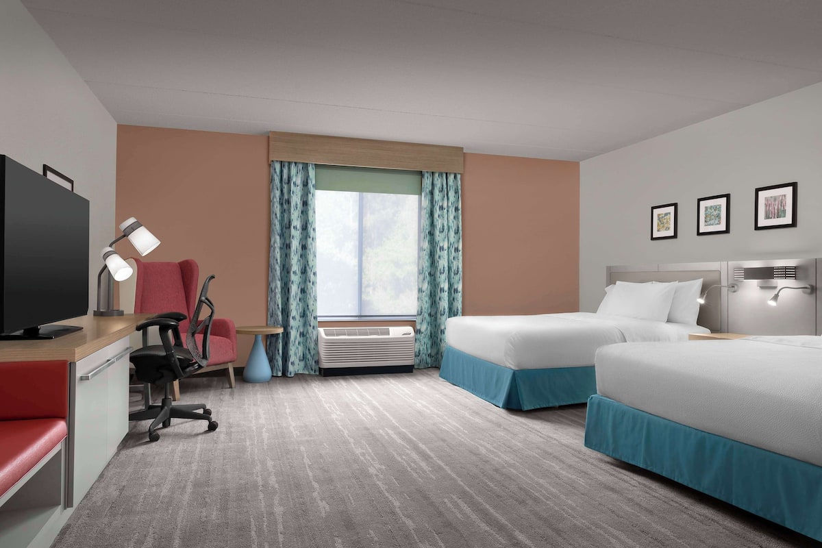 A hotel room with a desk, chair, two beds, a TV, air conditioning, and a window with curtains.
