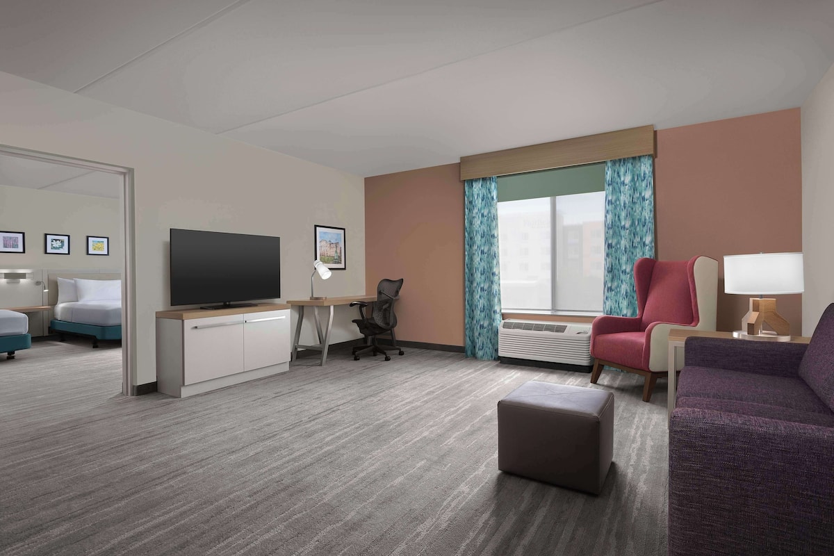 A hotel room with a flat-screen TV, a desk, a red armchair, a purple sofa, and a small ottoman.