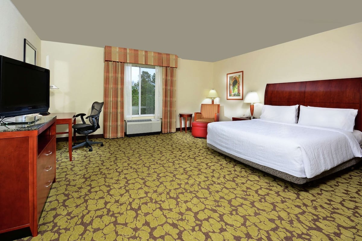 A hotel room with a bed, a desk with a TV, a chair, a window with curtains, a bedside table with lamps, and a carpeted floor.