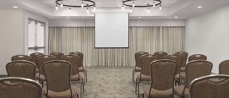 Une salle de conférence avec des rangées de chaises disposées face à un grand écran, une porte sur le côté gauche et un tapis à motifs.