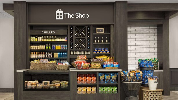 A store with a 'The Shop' sign, featuring chilled beverages, snacks, and packaged goods.