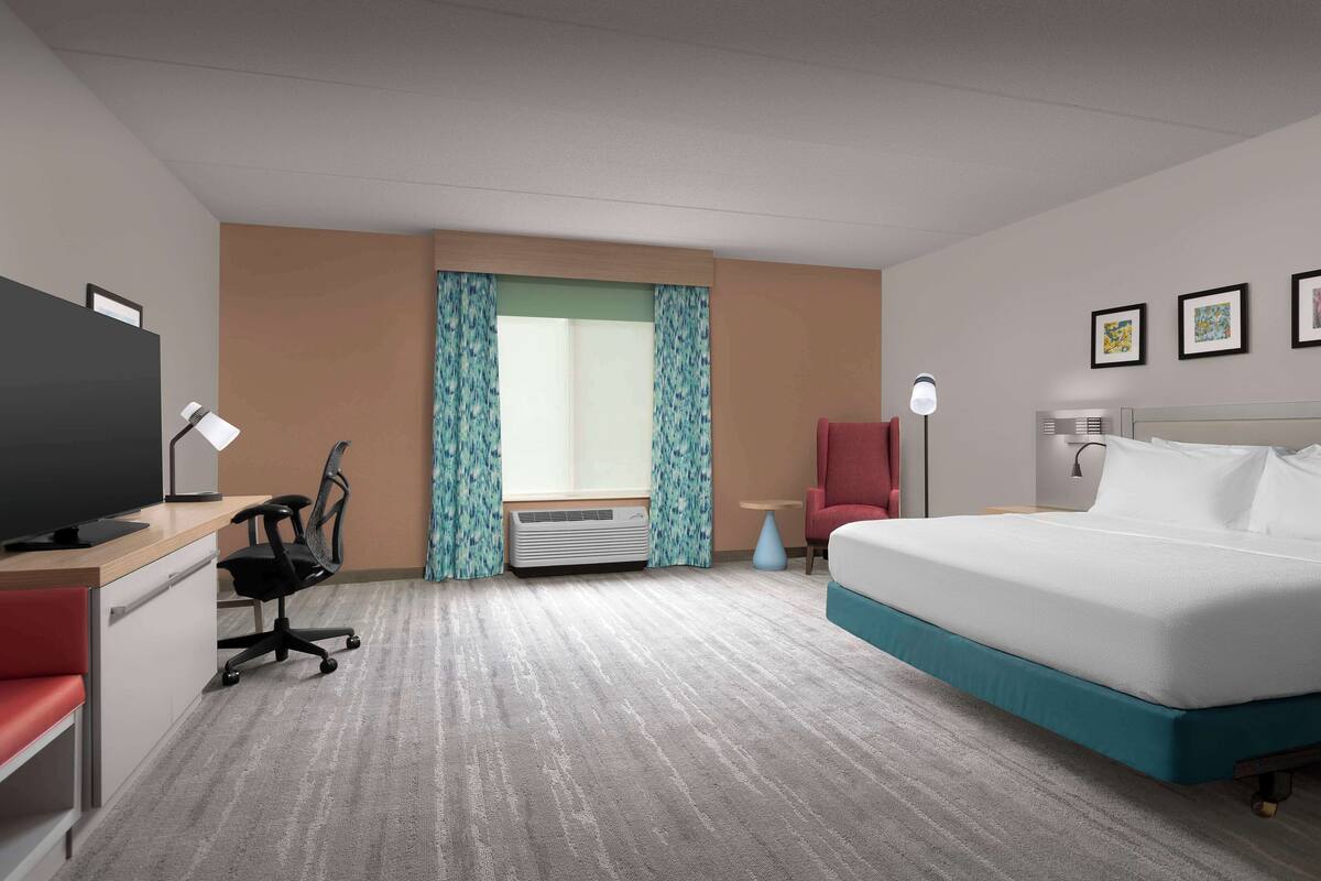 A hotel room with a bed, desk, chair, TV, air conditioning unit, and curtains.