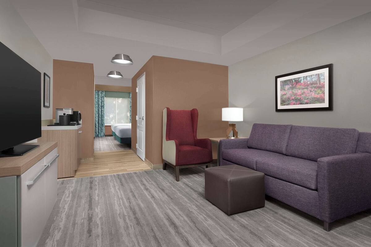 A hotel room with a sofa, armchair, ottoman, TV, coffee maker, and a framed picture on the wall.