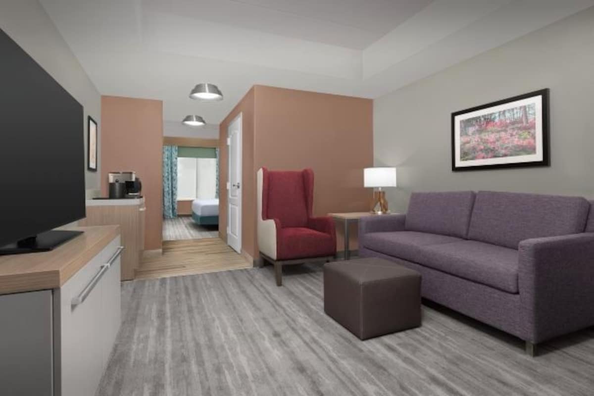 A hotel room with a television, a sofa, a red chair, a small table with a lamp, and a framed picture on the wall.