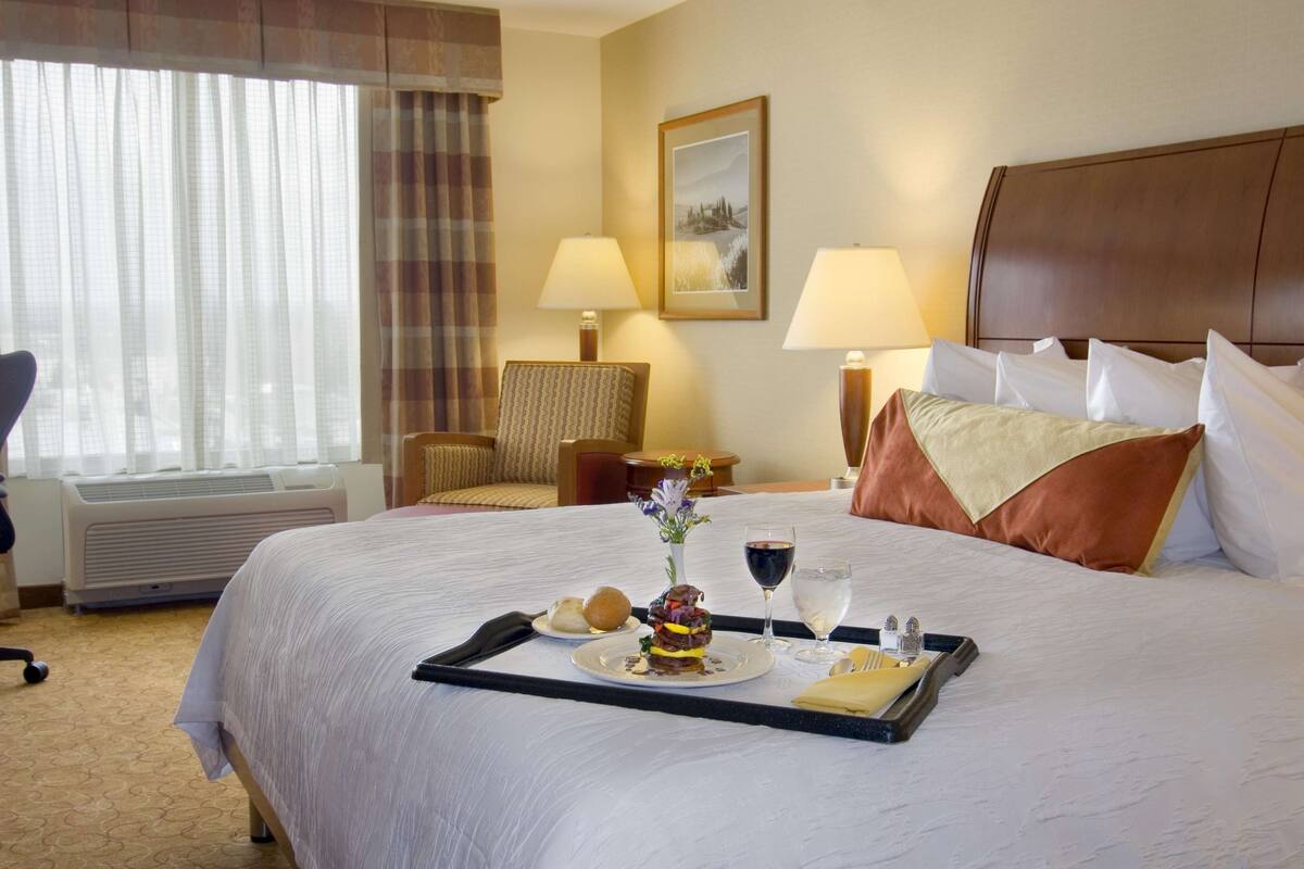 A hotel room with a neatly made bed, a dining table with a tray of food, a chair, and a lamp.