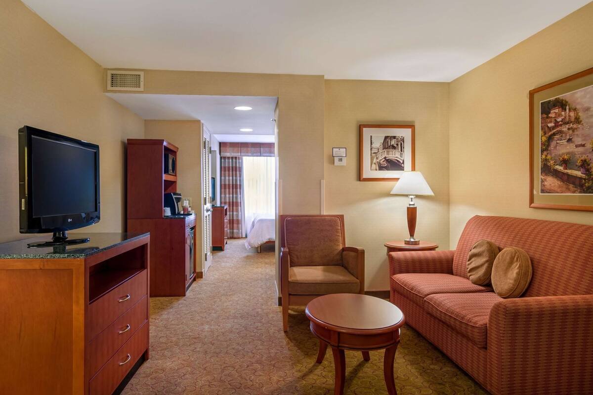 A hotel room with a television, a sofa, a chair, a small table, a lamp, and a painting on the wall.