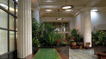 A lobby with a green-tiled bench, wooden chairs, potted plants, and marble columns.