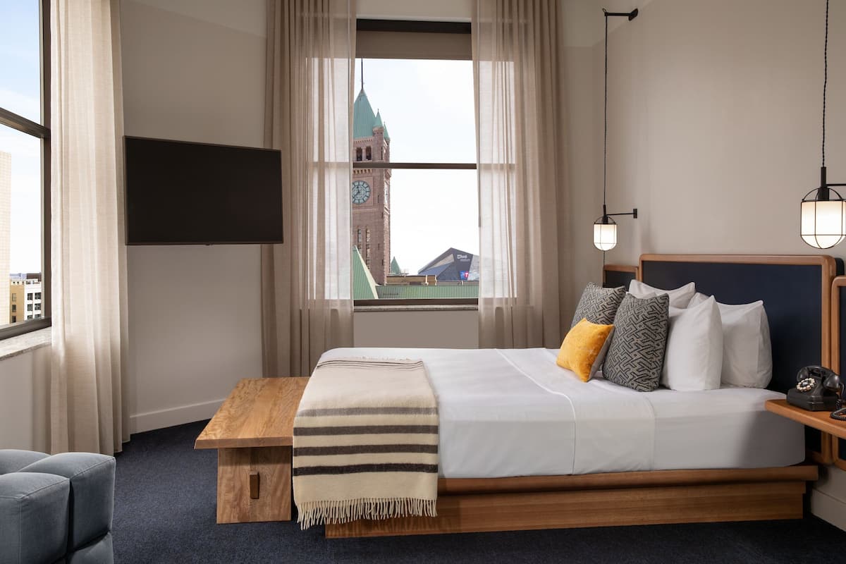 A modern hotel room with a large bed, a wooden bench, a TV mounted on the wall, and a view of a clock tower through the window.