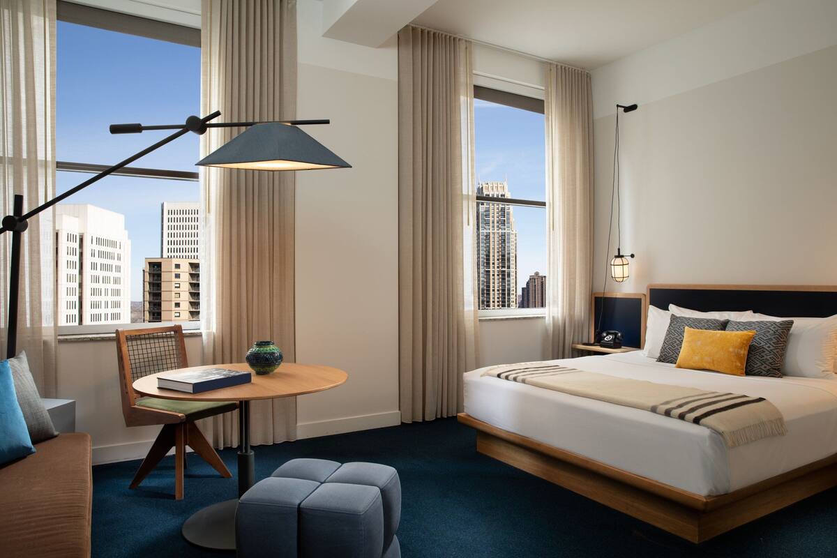 A hotel room with a large bed, a desk with a chair, a round table, and a view of tall buildings through large windows.