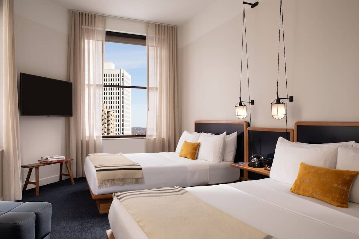 A hotel room with two beds, a TV, a desk, and a view of buildings.
