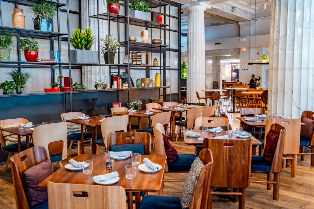 A restaurant with wooden tables and chairs, shelves with plants and books, and a marble column.
