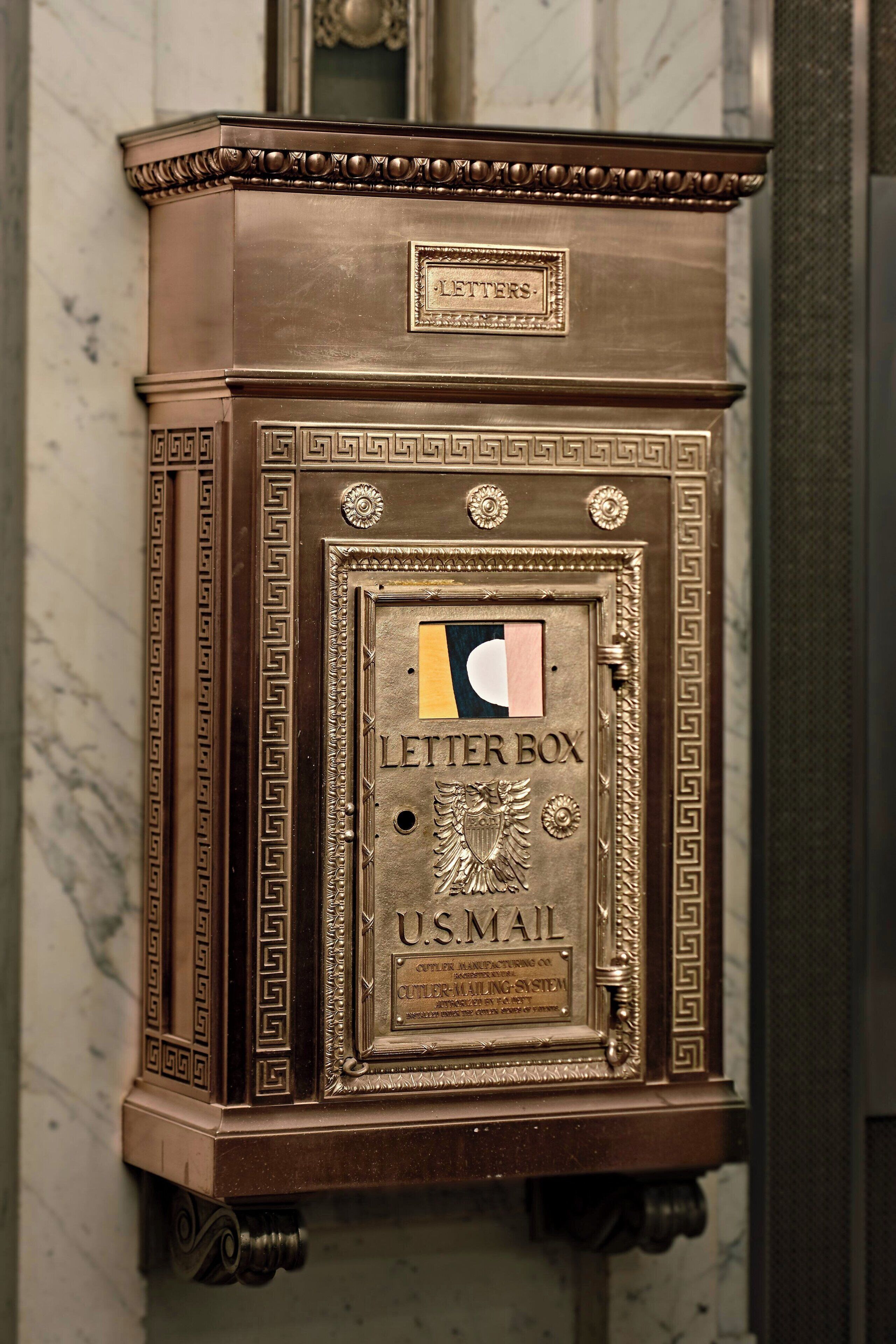 Caixa de correio antiga dos Correios dos EUA, com detalhes metálicos ornamentados e uma etiqueta com a inscrição “LETTER BOX U.S.MAIL”.