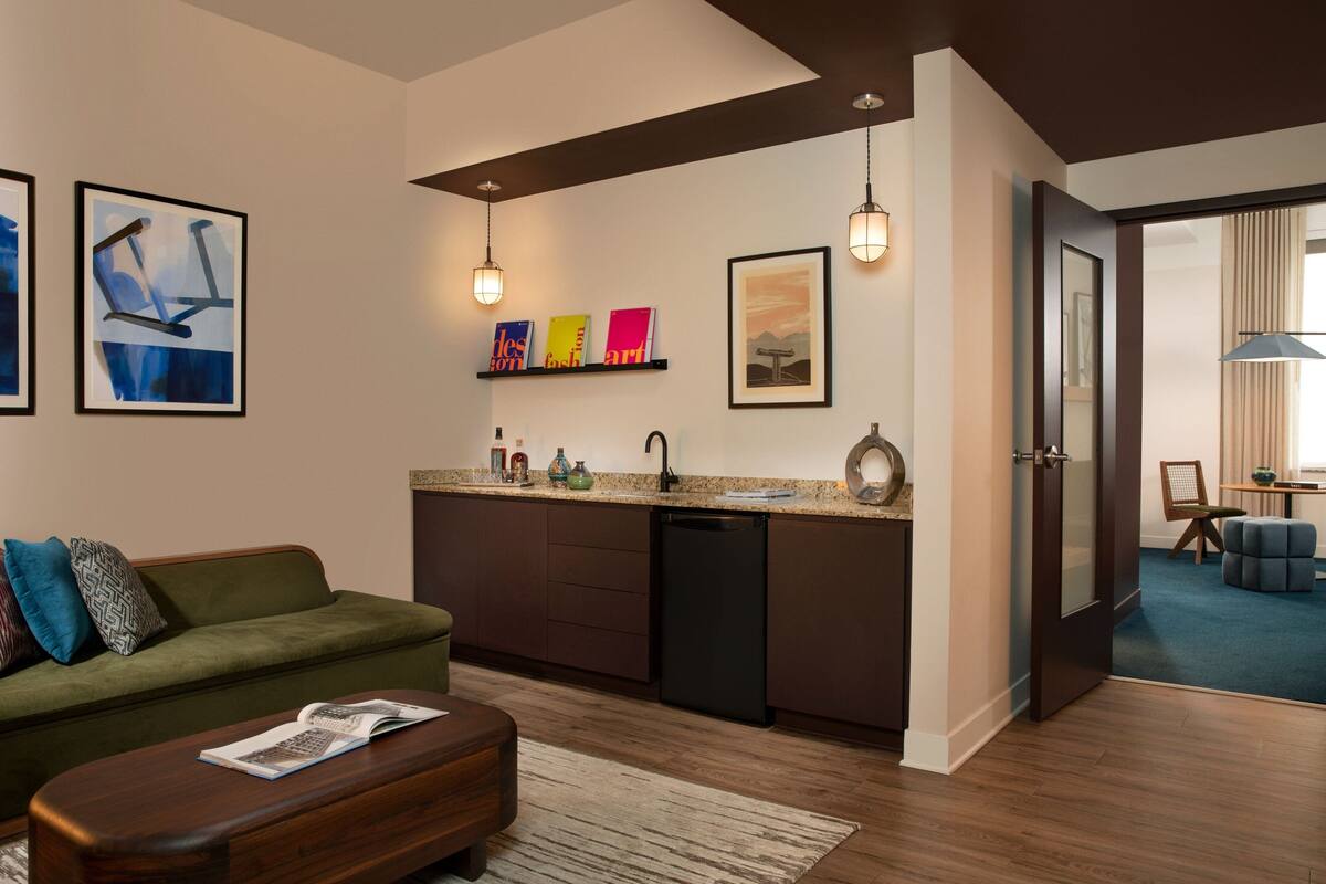 A modern living room with a green sofa, wooden coffee table, and a kitchen area with a dishwasher and countertop.