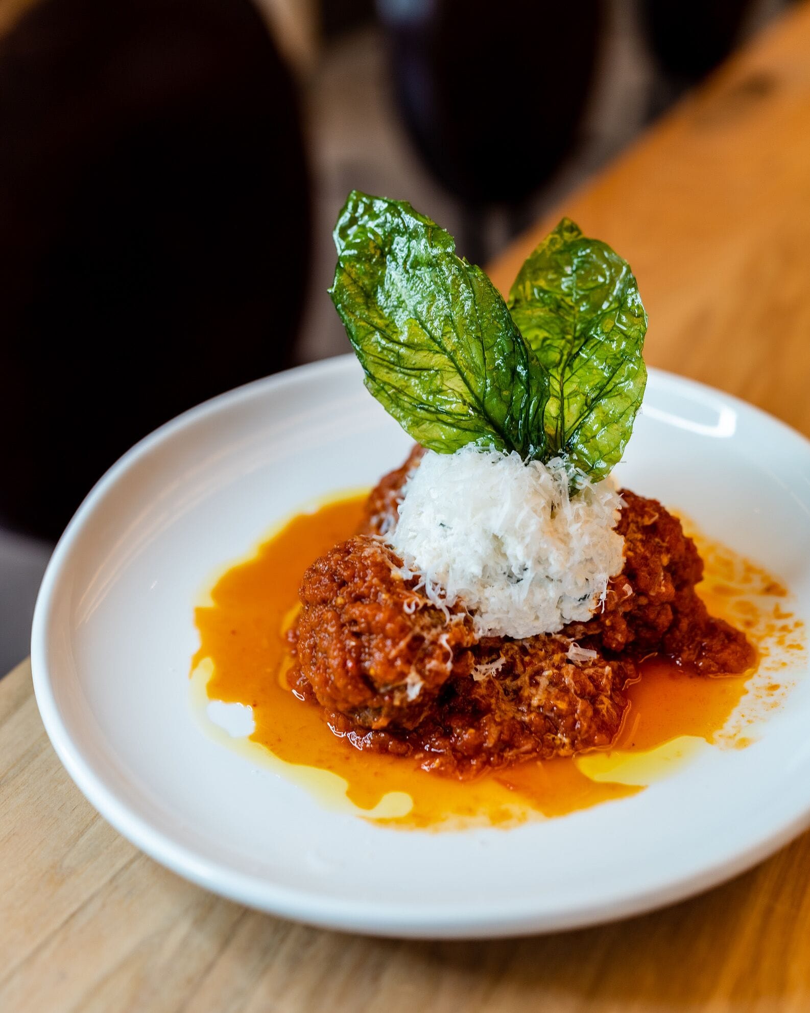 A white plate with a dish of meatballs in tomato sauce, topped with shredded coconut and a basil leaf.