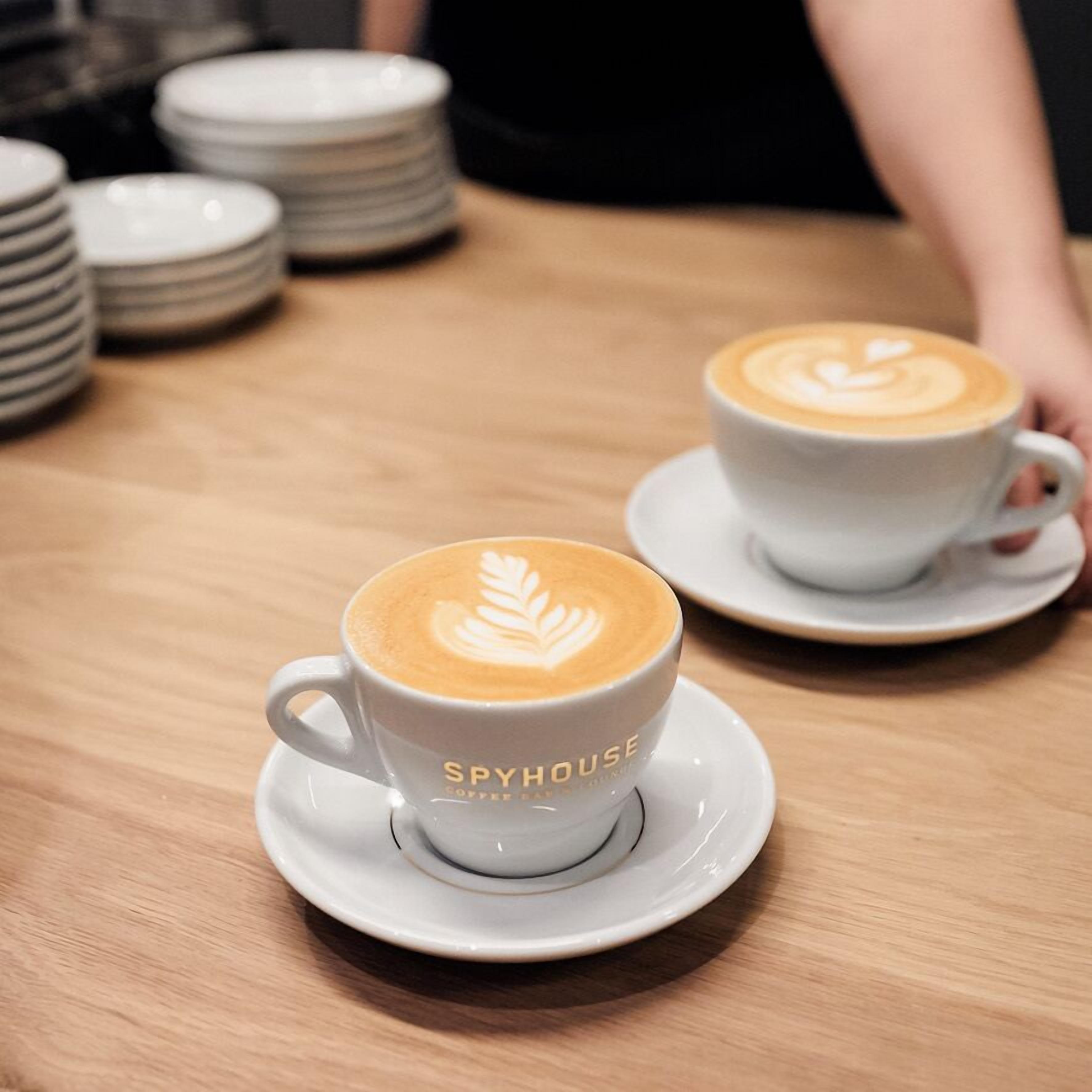 Two white cups of coffee with latte art on a wooden table, labeled "SPYHOUSE."