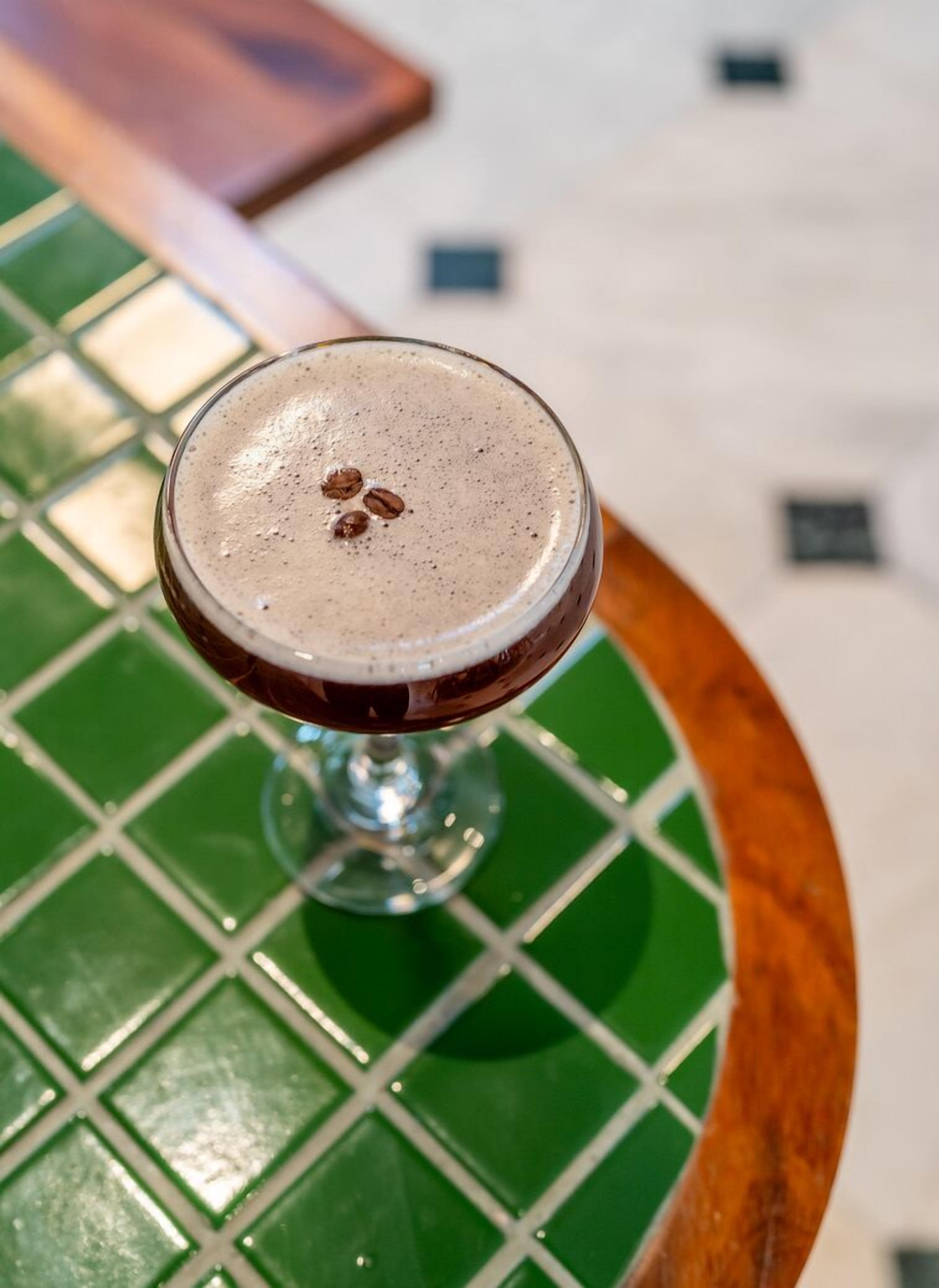 A glass of dark beverage with foam on top, placed on a green and white tiled table.