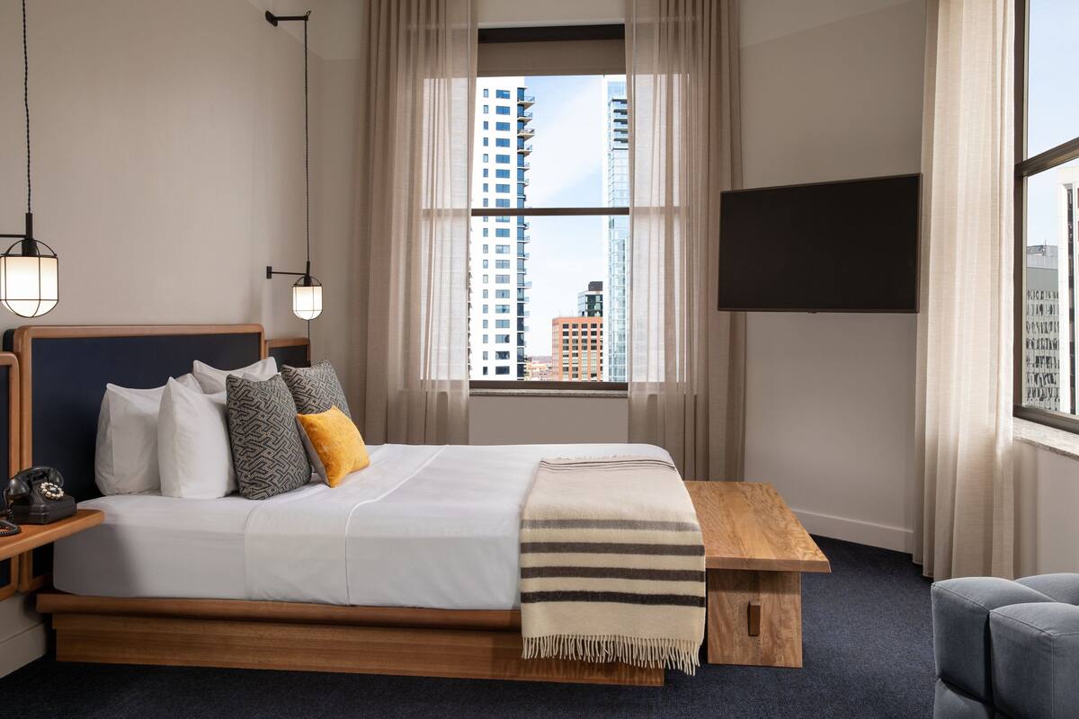 A modern hotel room with a large bed, a wooden bedside table, a bedside lamp, a TV mounted on the wall, and a view of the cityscape through the window.