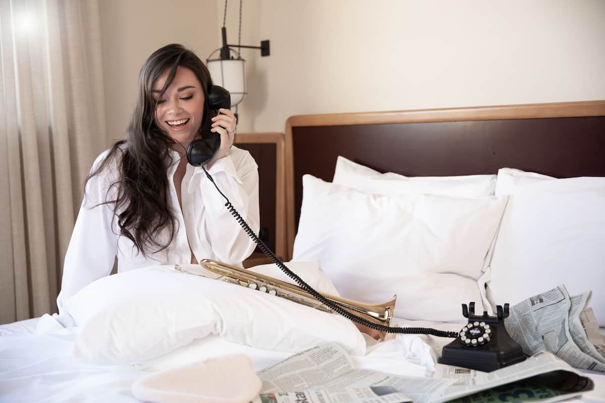 A hotel room with a bed, a telephone, a trumpet, and newspapers.