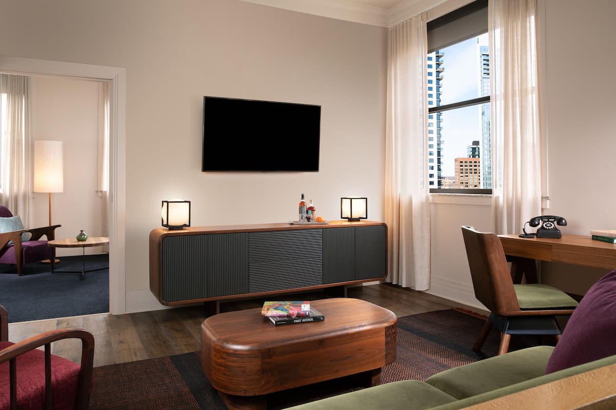 A modern hotel room with a flat-screen TV mounted on the wall, a wooden console table with a lamp, a small table with a lamp, a sofa, and a chair.
