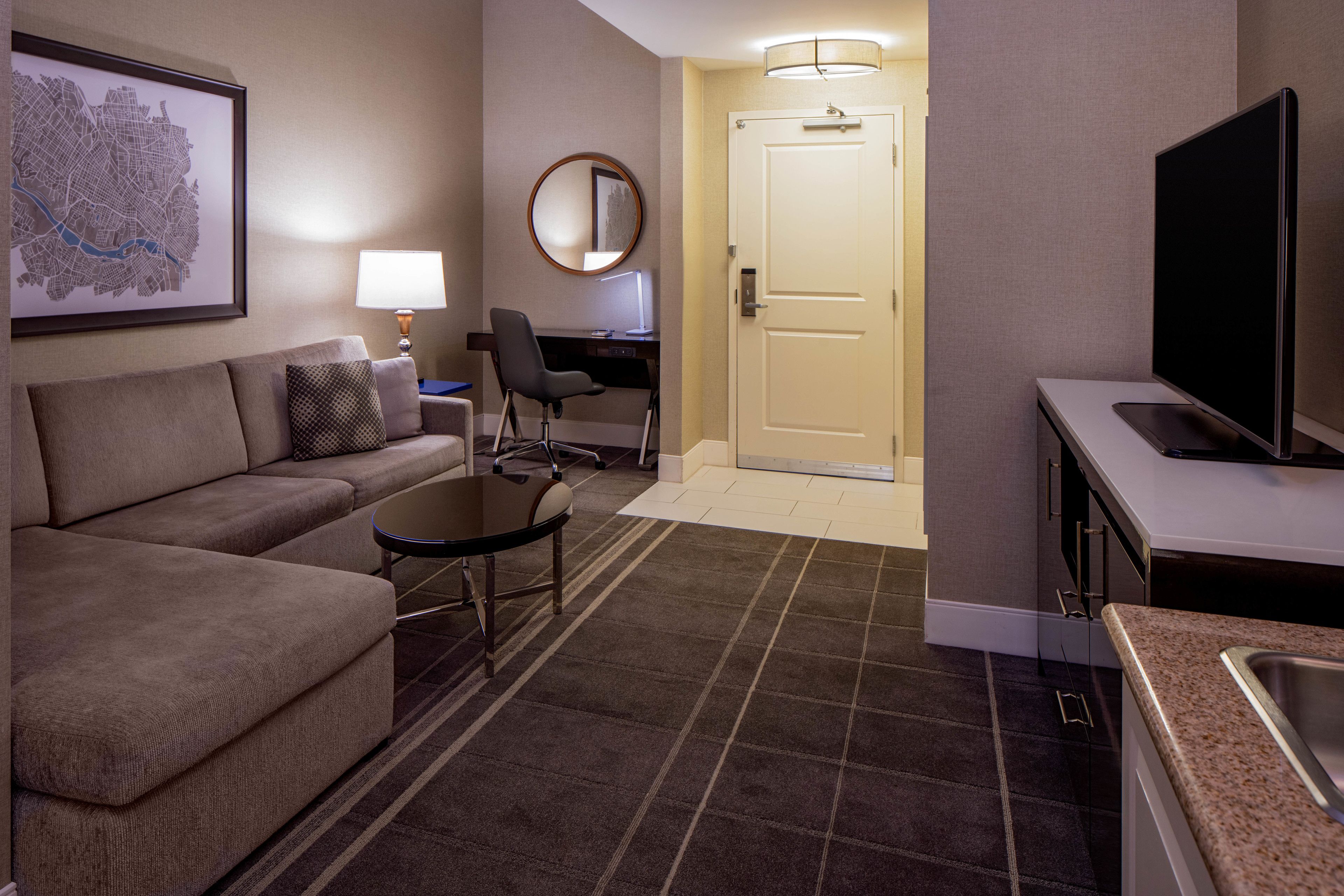 A hotel room with a sofa, a small table, a desk with a chair, a TV, and a door leading to another room.