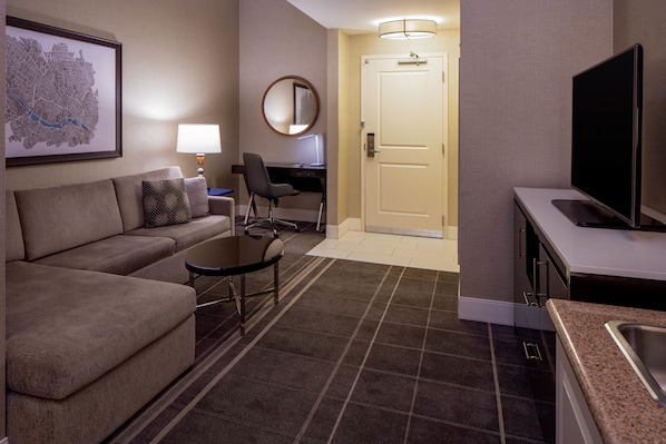 Habitación de hotel con sofá, una mesita, escritorio con silla, televisor y puerta que da a otra habitación.