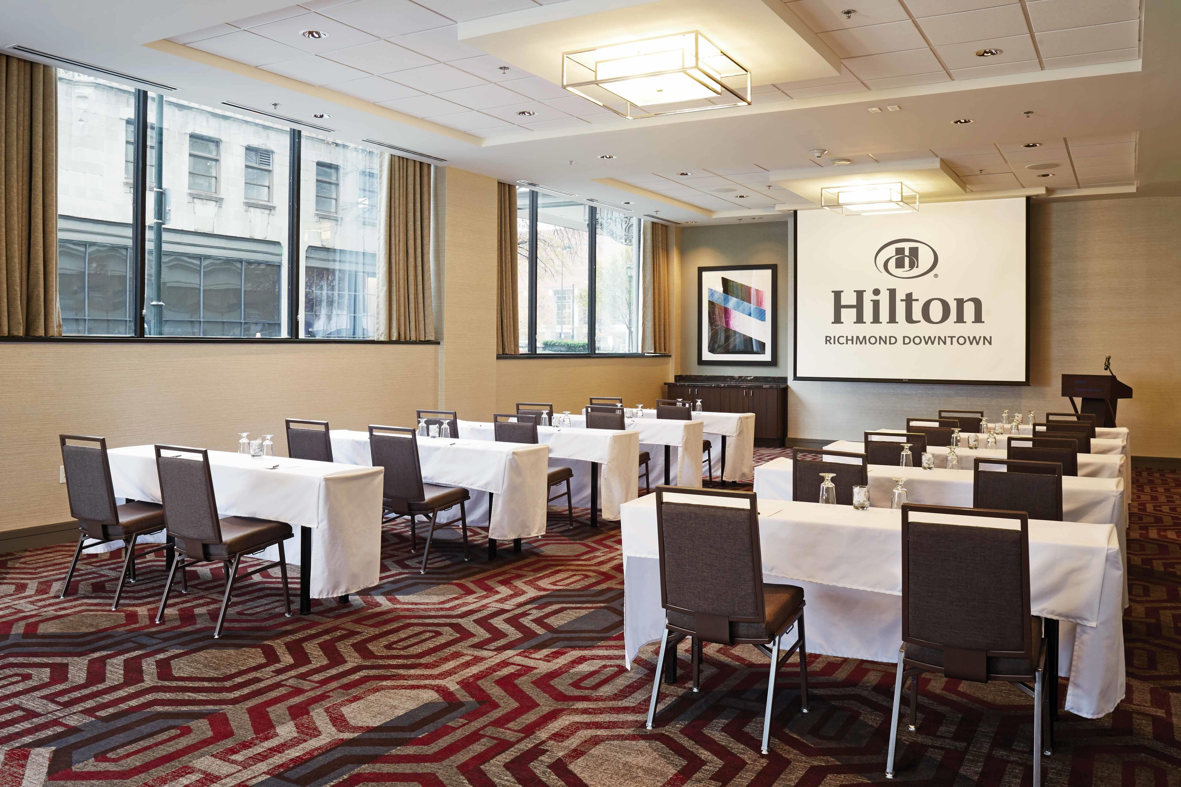 Une salle de conférence dotée de plusieurs tables et chaises, d’un grand écran affichant le logo de Hilton, et de grandes fenêtres équipées de rideaux.