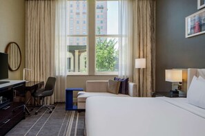 A hotel room with a bed, a desk with a chair, a sofa, and a window with curtains.