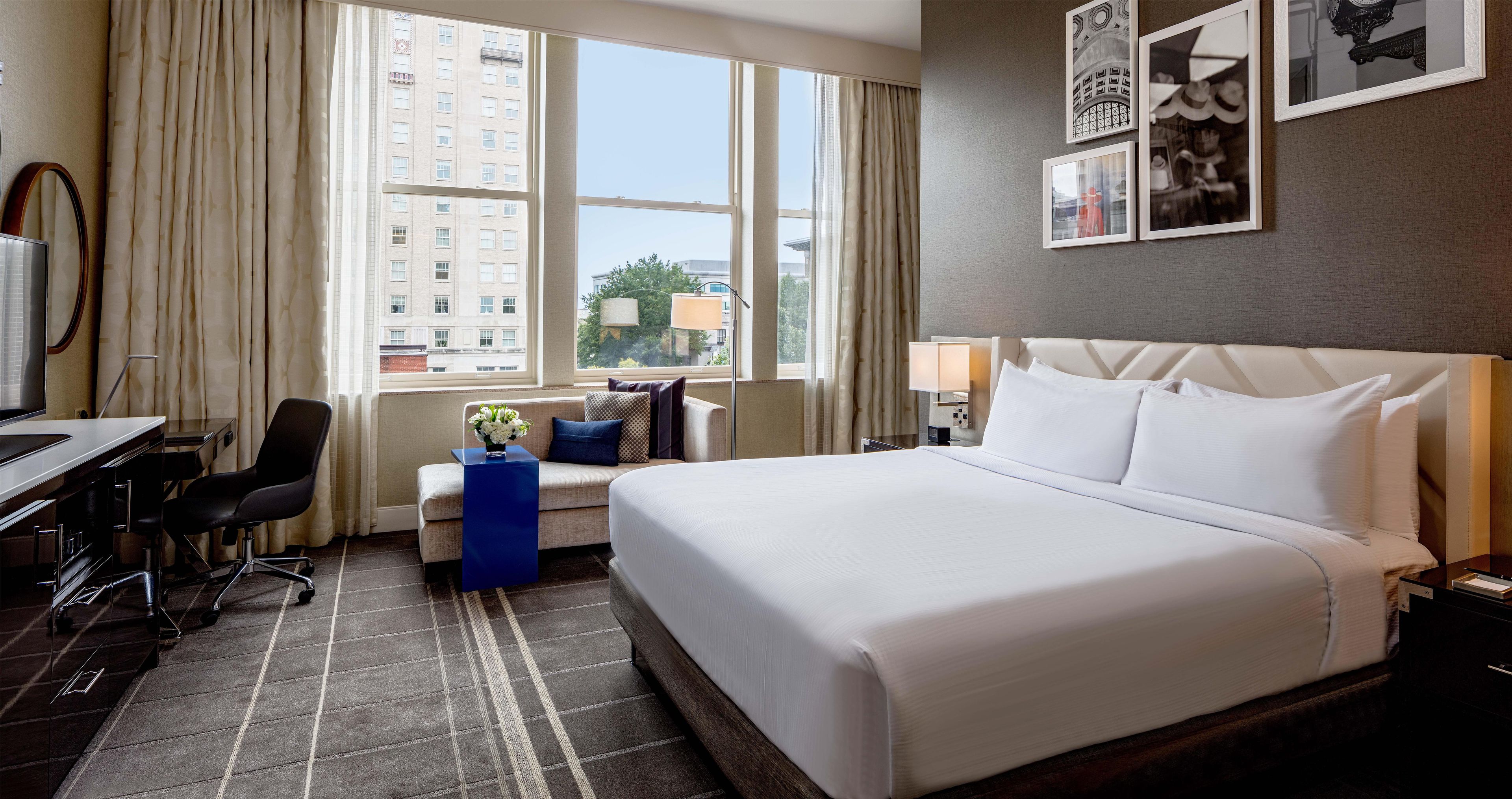 Habitación de hotel con una cama grande, un escritorio, una silla, una mesita con un florero y vistas a edificios a través de la ventana.
