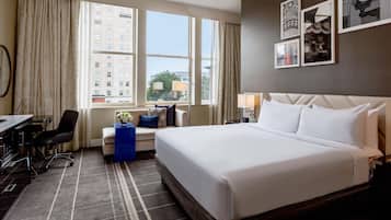 Habitación de hotel con una cama grande, un escritorio, una silla, una mesita con un florero y vistas a edificios a través de la ventana.
