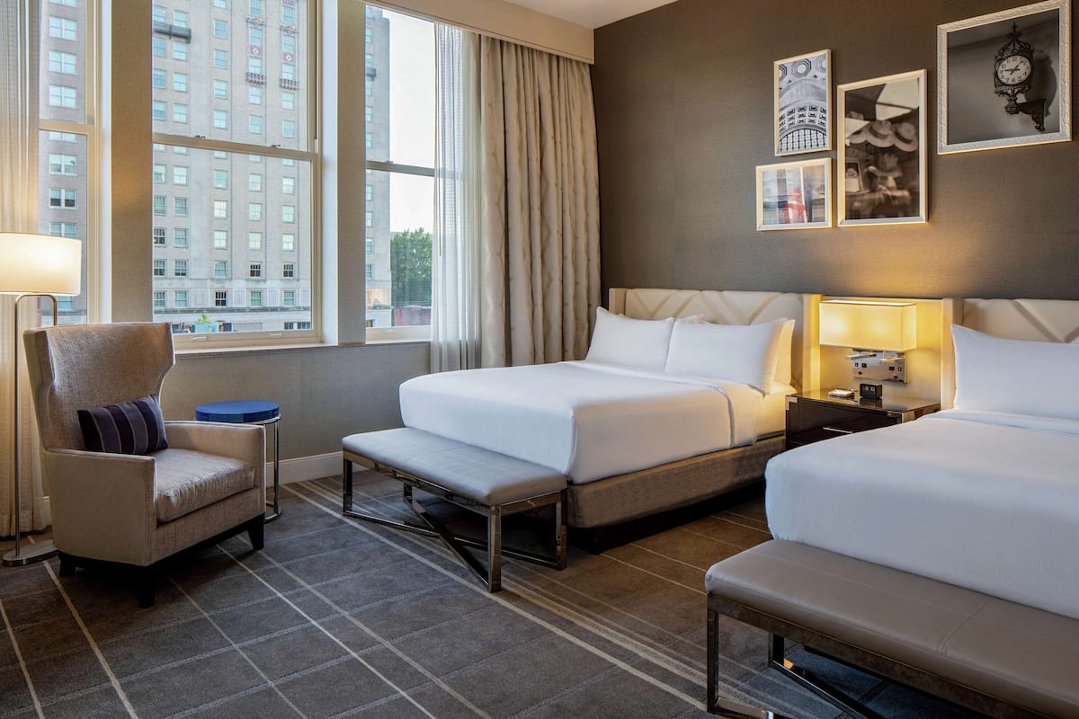 A hotel room with two beds, a chair, a bench, a nightstand, and a window with a view of a building.