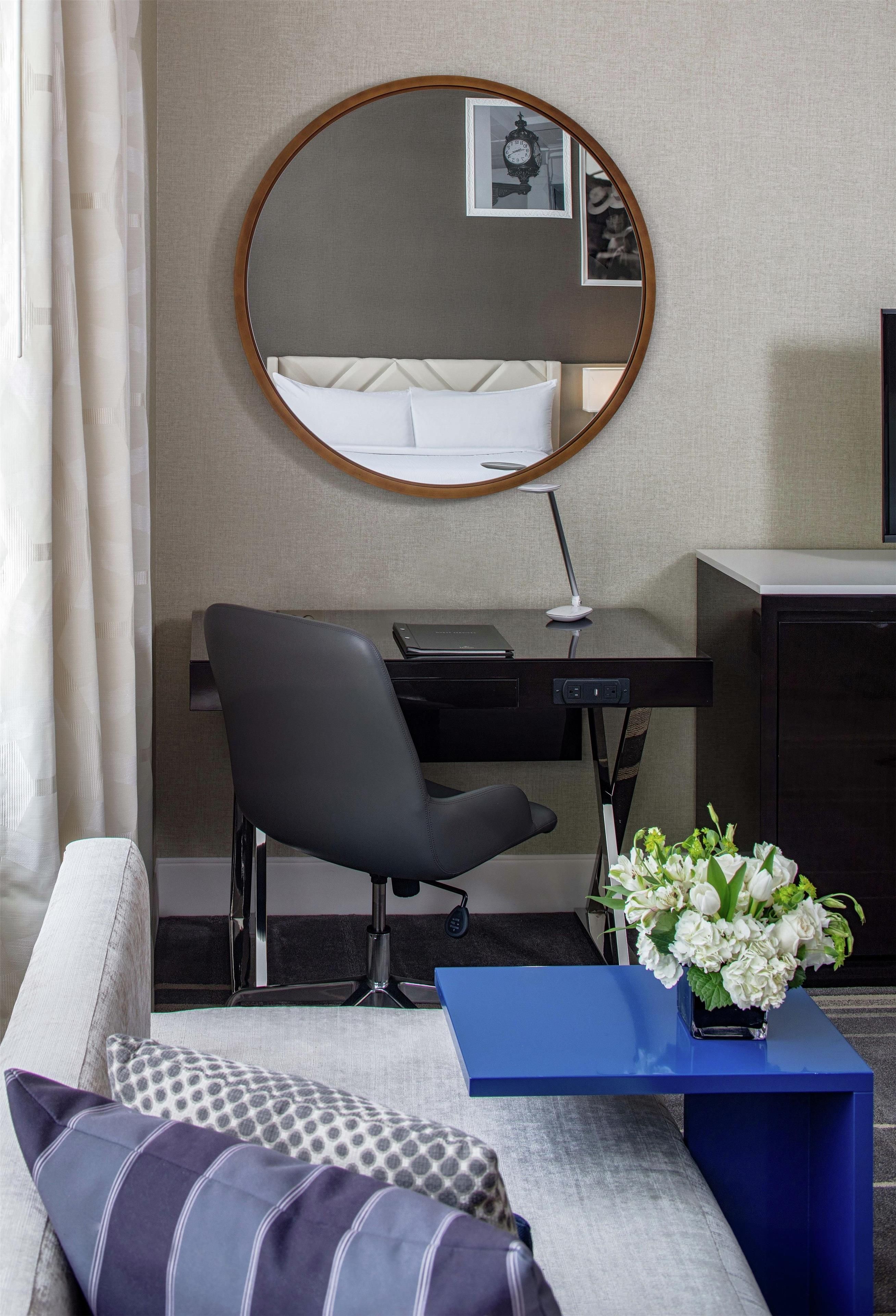 Quarto de hotel com espelho redondo, escrivaninha, cadeira, mesa lateral azul, vaso com flores e sofá com almofadas estampadas.