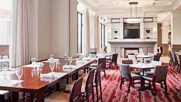 A hotel dining area with multiple tables set for dining, a television mounted above a fireplace, and large windows allowing natural light.