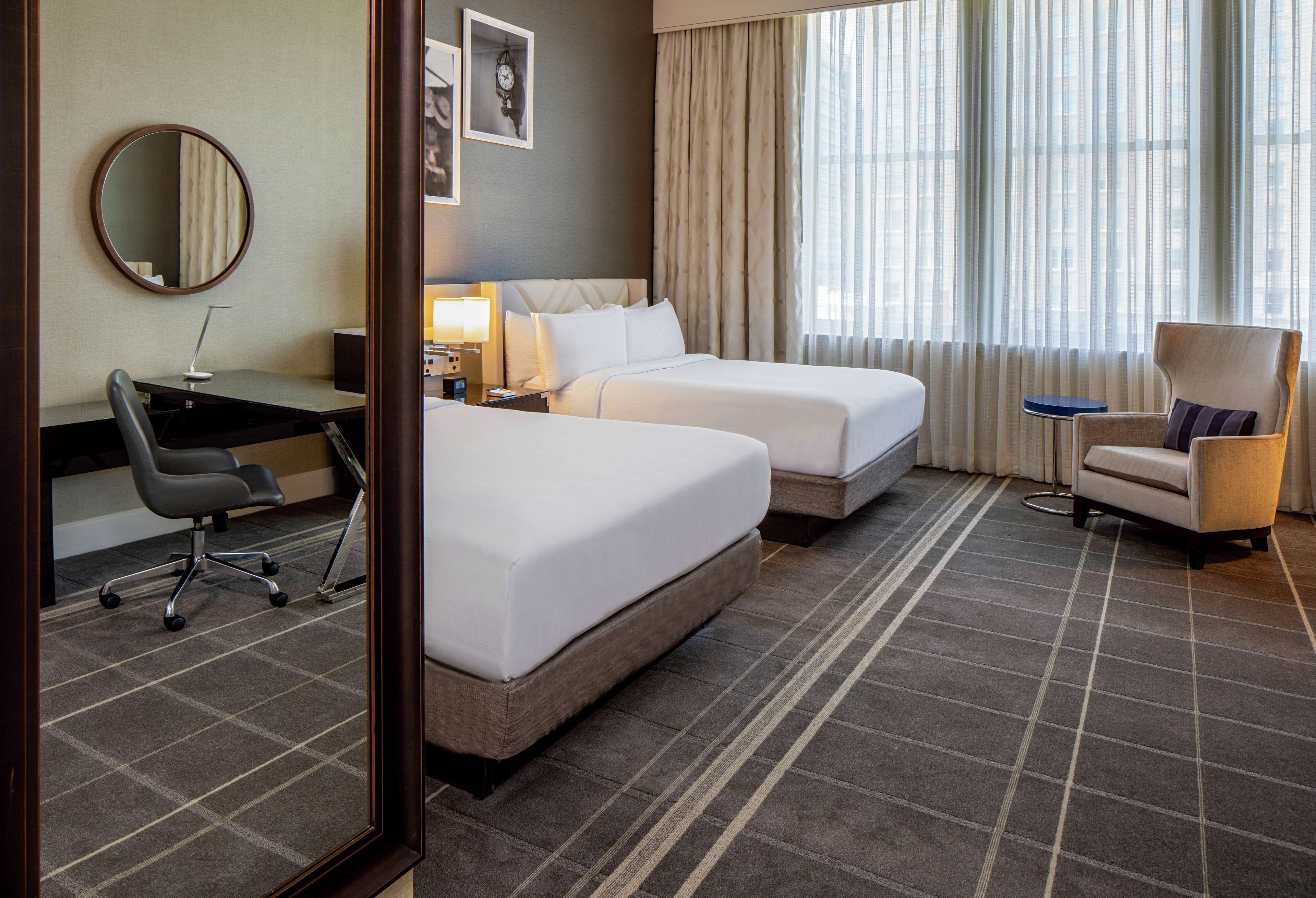 Quarto de hotel com duas camas, uma escrivaninha, uma cadeira, um espelho e uma janela com cortinas.