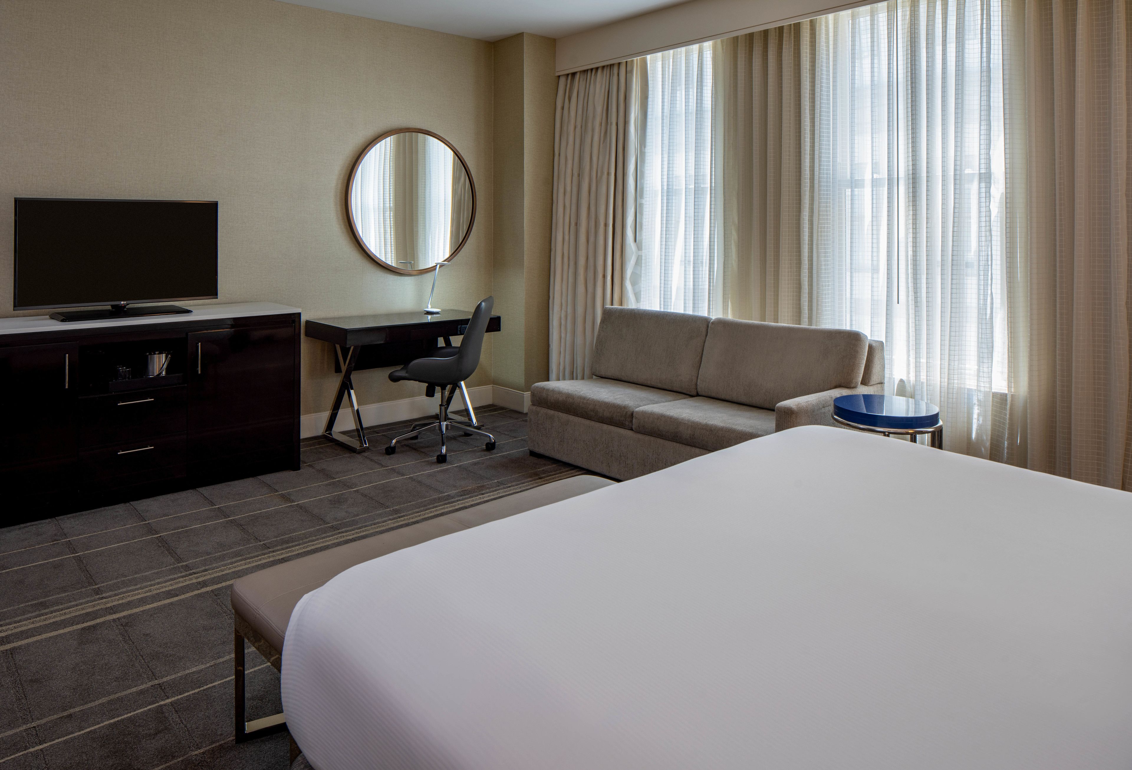 Une chambre d’hôtel avec un grand lit, un bureau avec une chaise, une télévision, un miroir rond et un canapé.