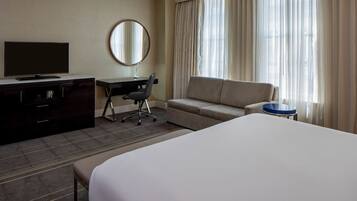 Une chambre d’hôtel avec un grand lit, un bureau avec une chaise, une télévision, un miroir rond et un canapé.