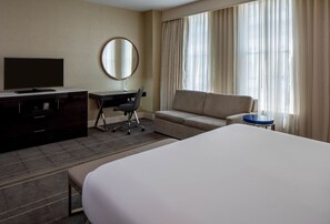 Una camera d'albergo con un letto grande, una scrivania con una sedia, una televisione, uno specchio rotondo e un divano.